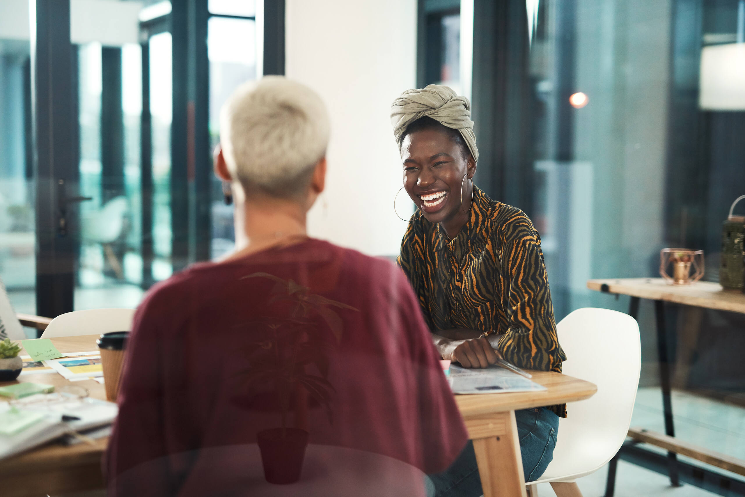 Two women, one Black wearing a headwrap and patterned top, one with short white hair, laughing together at a table in a bright office setting, depicting positive recovery support