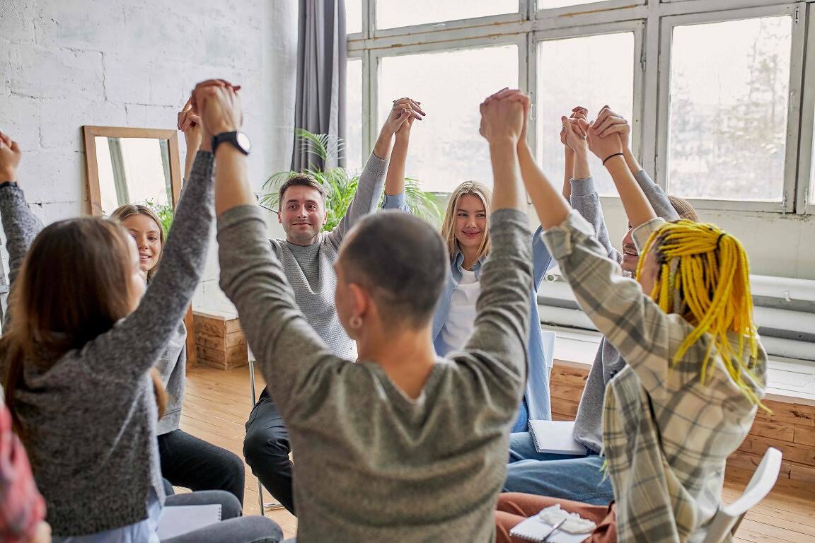 Diverse group of people in a therapy circle raising joined hands, showing unity and support in addiction recovery