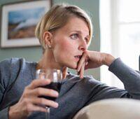 Concerned young blonde woman in gray sweater sitting alone, holding a glass of red wine while looking distressed and contemplative, suggesting alcohol dependency issues