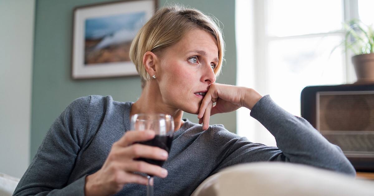 Concerned young blonde woman in gray sweater sitting alone, holding a glass of red wine while looking distressed and contemplative, suggesting alcohol dependency issues