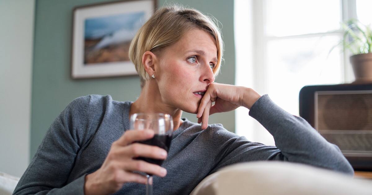 Concerned woman holding glass of red wine, looking pensive in home setting, possible signs of alcohol dependency