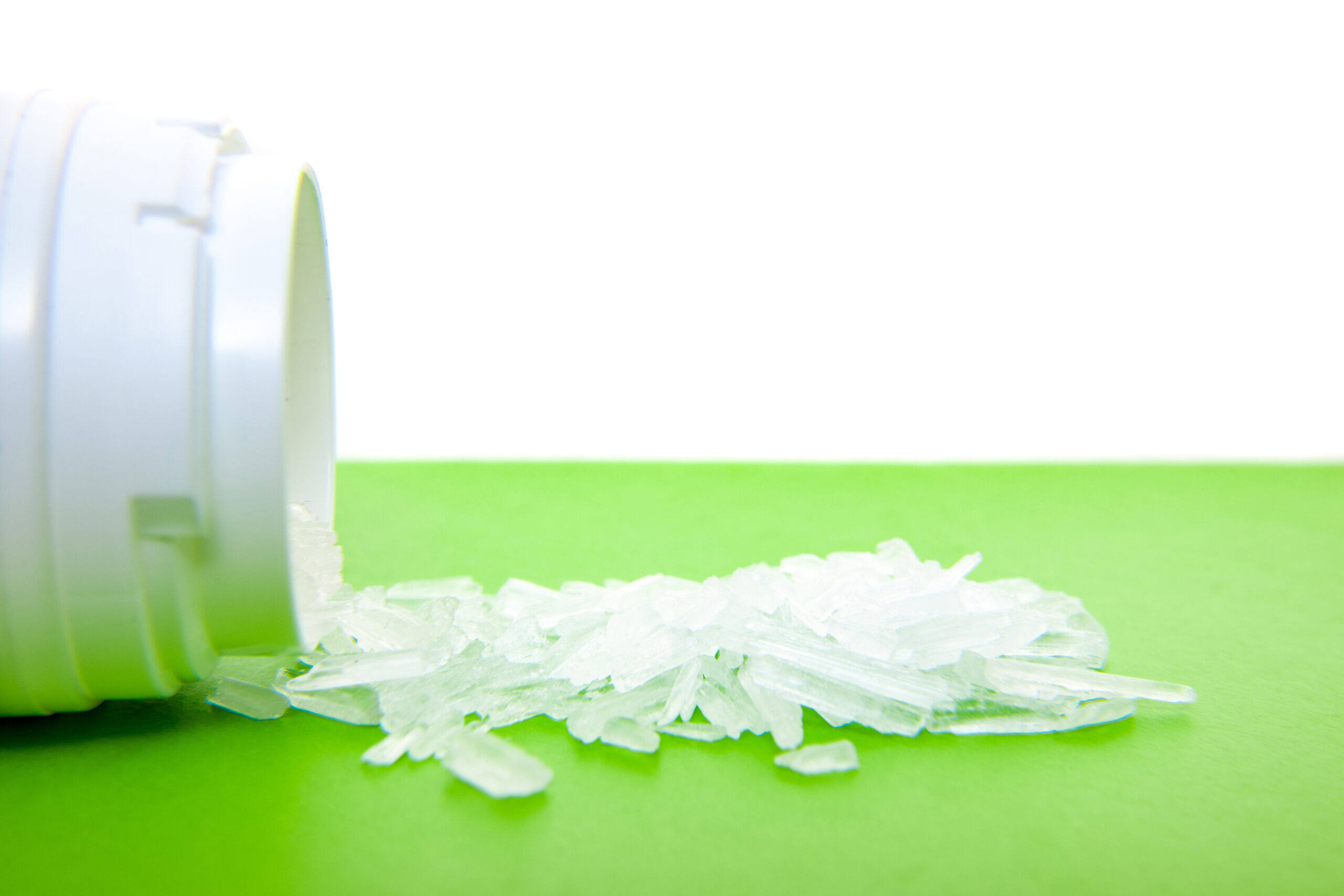 Close-up of crystal methamphetamine shards spilled from a small container on a bright green surface, illustrating the dangers of ice addiction