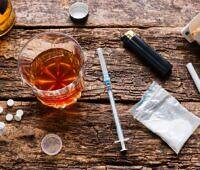 Assortment of drugs and alcohol on a wooden surface, including a glass of whiskey, syringe, white powder, pills, cigarettes, and lighter, illustrating the dangerous combination of cocaine and alcohol