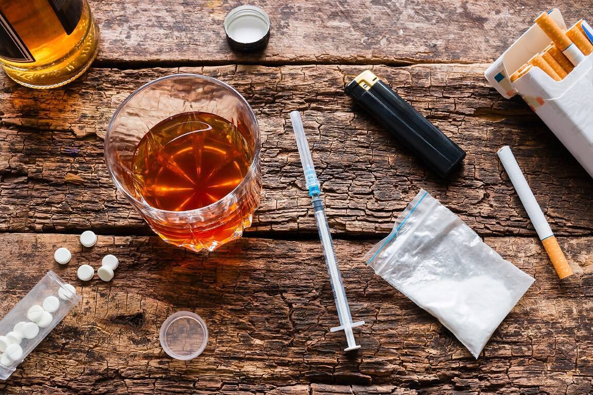 Assortment of drugs and alcohol on a wooden surface, including a glass of whiskey, syringe, white powder, pills, cigarettes, and lighter, illustrating the dangerous combination of cocaine and alcohol