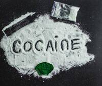 White powder spelling out 'COCAINE' on a black surface, with a small plastic bag and a green leaf, illustrating the illicit nature and health risks of cocaine use