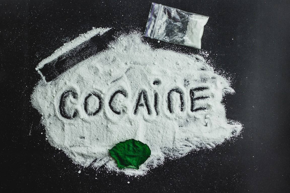 White powder spelling out 'COCAINE' on a black surface, with a small plastic bag and a green leaf, illustrating the illicit nature and health risks of cocaine use