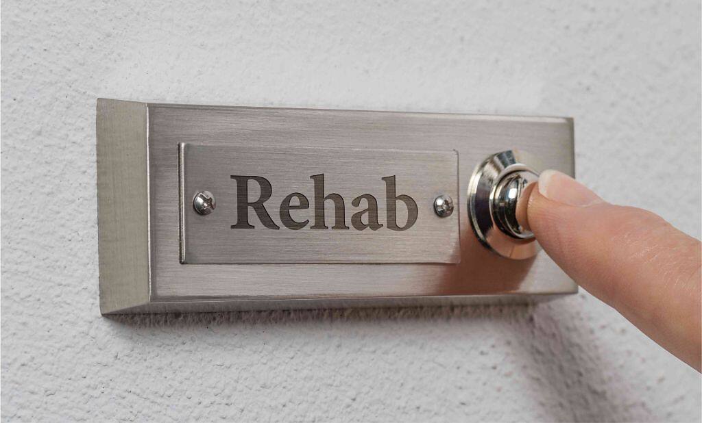 Close-up of a finger pressing a doorbell labeled 'Rehab', symbolizing the first step toward recovery