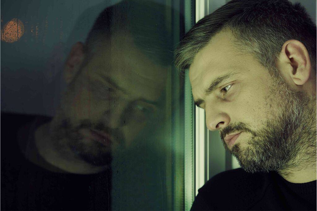 A man looking contemplative, leaning against a window, symbolizing introspection during addiction recovery.