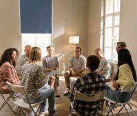 group of people in a rehab group therapy meeting in a large room with many window sitting in a circle on chairs listening to one woman talking