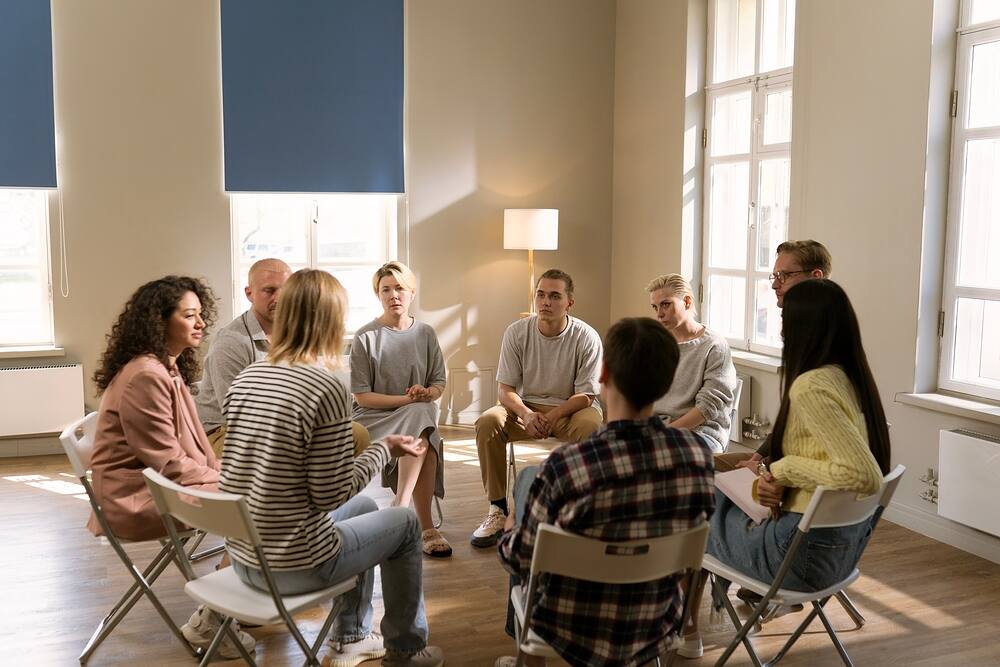 group of people in a rehab group therapy meeting in a large room with many window sitting in a circle on chairs listening to one woman talking