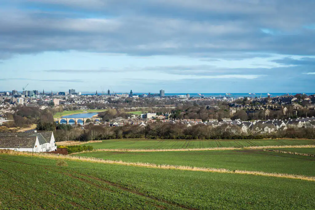 Panoramic view of Aberdeen city skyline with countryside fields in foreground, showcasing urban and rural Scotland|Serene beach scene in Aberdeenshire with a small boat on sand, rocky shoreline, and calm sea under a dramatic sky, symbolizing a journey towards healing and sobriety|Female therapist in green sweater listening attentively to patient during addiction counseling session, holding document|Young woman with curly hair in beige jacket engaged in conversation during an addiction therapy session, showing attentive listening|Happy family of four smiling outdoors, parents giving piggyback rides to two children, representing successful recovery and family support after addiction rehabilitation|Young man in pink shirt engaging in conversation with older male therapist taking notes, surrounded by other group members in a bright, modern therapy room|Picturesque ivy-covered stone manor house in Aberdeenshire with dormer windows, surrounded by trees and featuring a beautiful lavender garden in the foreground, ideal for a peaceful rehabilitation setting