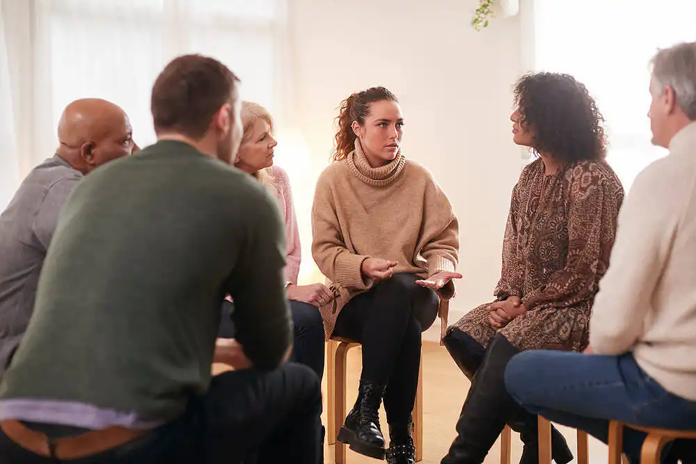 Diverse group of people sitting in a circle during a therapy session, focused on a woman speaking in the center|Woman drinking coffee while participating in a virtual group therapy session on her computer, showcasing online addiction recovery support during social distancing