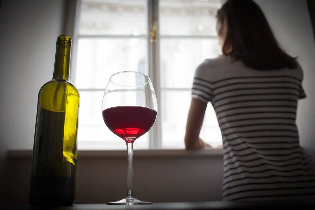 Woman looking out window with wine bottle and glass in foreground, depicting alcohol addiction and isolation