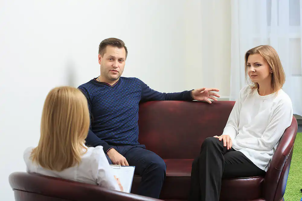 A Man and woman sitting on a couch. A third person sits on a chair facing them taking notes suggesting she is their therapist