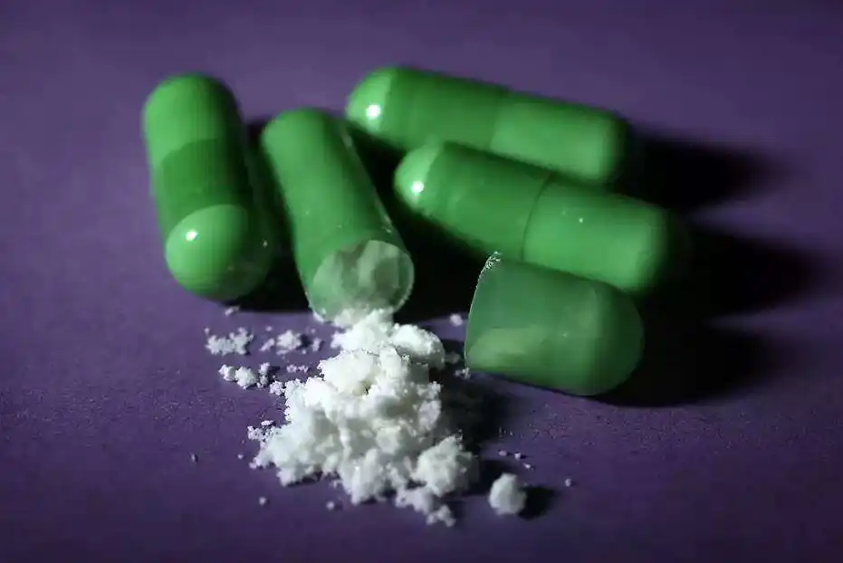 Several green drug capsules next to a small pile of white powdery substance on a purple background