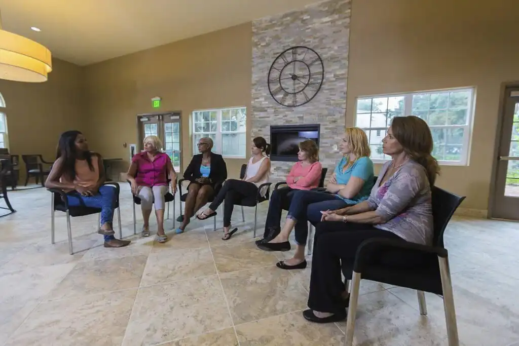 Diverse group of women participating in a therapy session at a rehabilitation centre, sitting in a circle in a modern, well-lit room with a large clock on the wall.