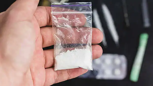 Close-up of a hand holding a small plastic bag containing white powder, ketamine, with drug paraphernalia visible in the background|Black and white image of a person walking towards a large keyhole-shaped opening in a concrete wall, symbolizing hope and the path to overcoming addiction|Silhouette image of two women, one holding a pill or substance to her mouth, the other with head in hand, depicting the emotional and physical toll of addiction|A diverse group of people sitting in a circle during a therapy or support group session, representing community-based addiction recovery programs