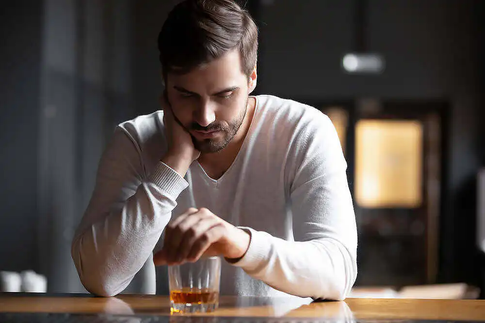 Troubled man leaning on counter, looking pensively at glass of alcohol, considering addiction treatment|Man holding up hand in refusal gesture towards a glass of whiskey, indicating potential alcohol dependency awareness