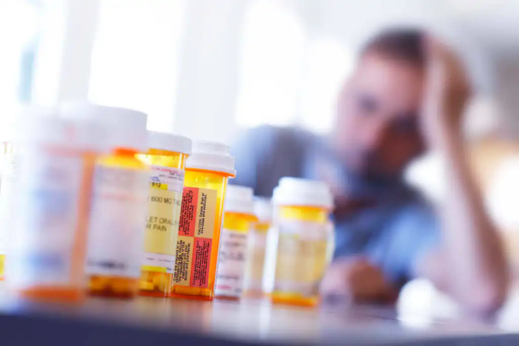 Multiple prescription medication bottles in foreground with blurred image of distressed person holding head in background, illustrating struggle with prescription drug dependence