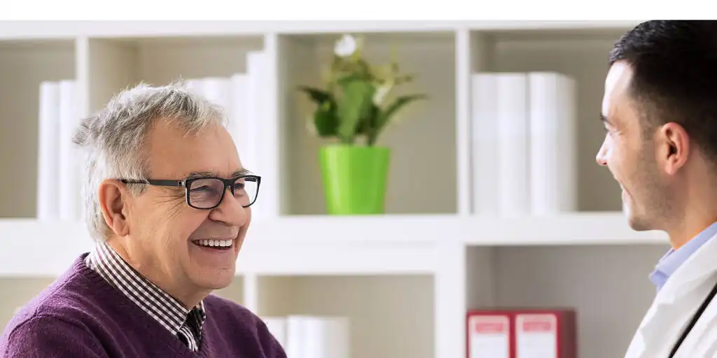Smiling elderly man in glasses speaking with a young doctor in a bright, modern medical office with bookshelves|Tranquil Hertfordshire scene with a clear stream flowing past quaint cottages and lush greenery, featuring a small wooden bridge and blooming flowers, symbolizing a peaceful journey towards healing and rehabilitation|Diverse group of people engaged in a positive therapy session, with a man in blue shirt speaking and others listening attentively, illustrating collaborative healing in addiction treatment|Collage showing luxurious living room, comfortable bedroom, and fitness area with clients exercising, representing the holistic approach to alcohol rehab in Hertfordshire