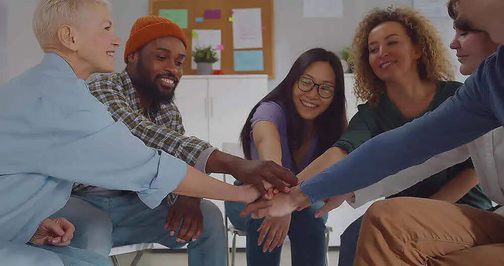 Diverse group of smiling people in a circle joining hands during a drug rehabilitation support group session, showcasing unity and mutual support|Two young women with visible signs of substance abuse, including bruises and disheveled appearance, highlighting the physical toll of drug addiction|||Diverse group of people sitting in a circle on yoga mats during a therapy or support group session, with a man gesturing as he speaks to the others in a bright, modern room with exposed brick walls|Close-up portrait of a cheerful middle-aged man with beard, wearing a blue shirt, smiling brightly, symbolizing success and happiness in addiction recovery|Group of diverse individuals in a supportive circle, joining hands in the center, smiling and engaged in a rehab or therapy setting, showcasing community and teamwork in recovery|Therapist in white clothing speaking with a male patient in a brown sweater during an individual therapy session, with colorful art therapy materials visible in the background|Close-up of two people holding hands in a supportive gesture, symbolizing empathy and connection during drug rehabilitation|Two people hugging tightly in a support group setting, with an older man with grey hair and beard embracing another person, while others look on supportively in the background|Collage of three images showing a brick residential rehab center exterior, a comfortable private bedroom, and a group therapy session with diverse participants