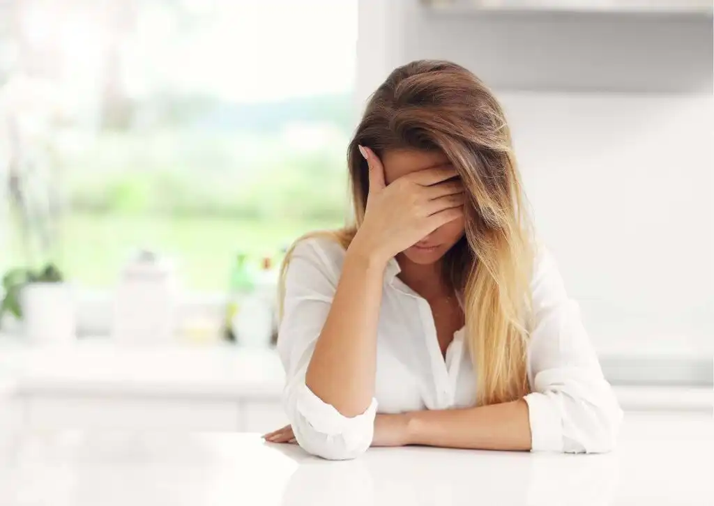 Distressed woman with hand covering face, possibly experiencing anxiety or discomfort from alcohol withdrawal|Young woman with blonde hair sitting at a table, covering her face with her hand, showing signs of distress or fatigue possibly related to substance abuse or withdrawal