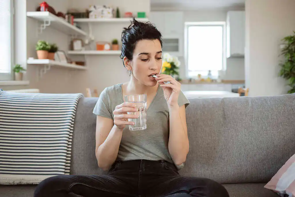 Young woman sitting on couch taking medication with glass of water in living room setting|Doctor consulting with a patient holding a beer bottle and pill container, addressing substance dependency issues.